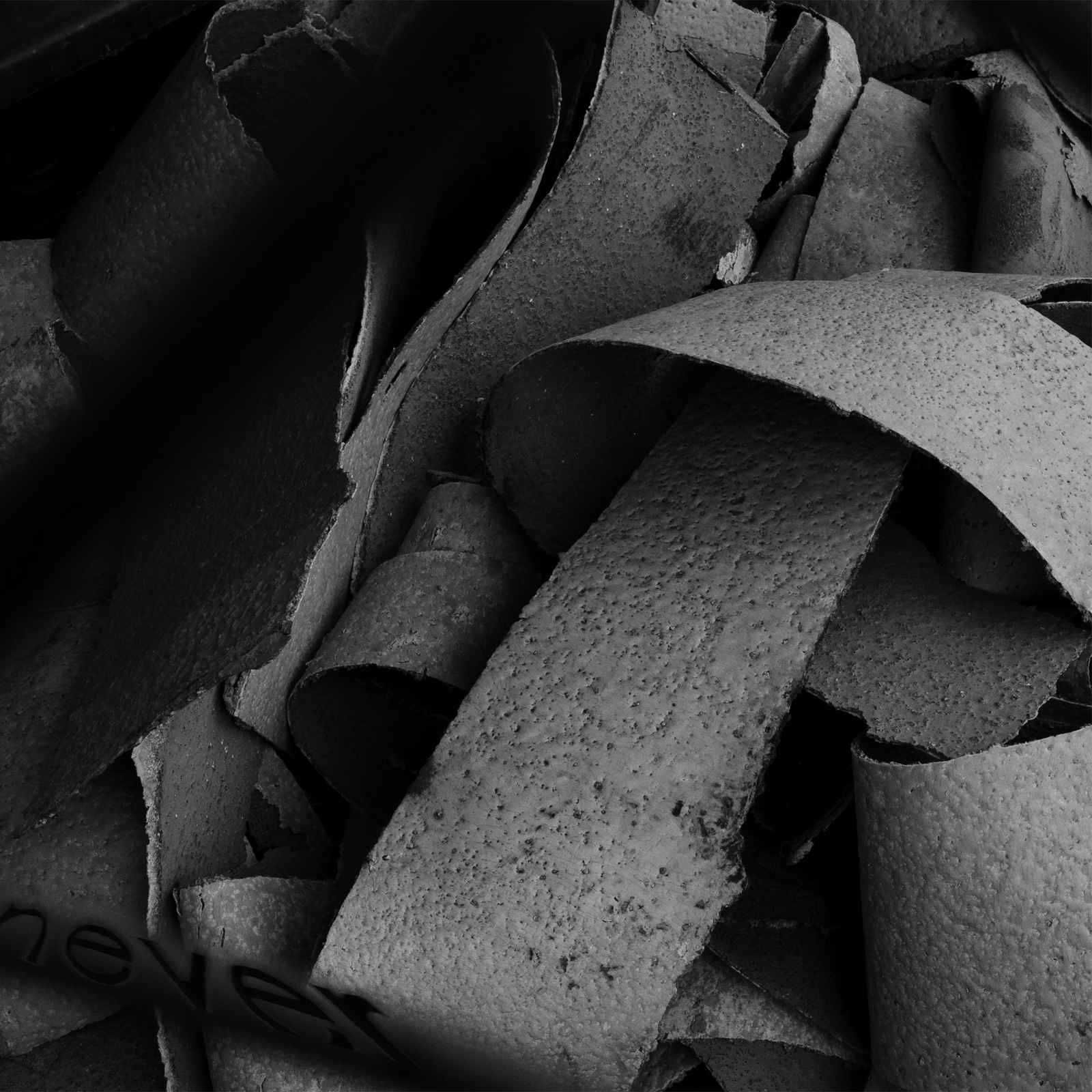Dark black-and-white abstract photograph of dirty, industrial strips of rubber, with the word “never” in a stretched black font above a black wire crossing the lower-left corner.